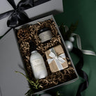 Luxurious Christmas gift box featuring Piotis Naturals' best-selling Calming Cream, Rosewater Hand & Body Lotion, and a beautifully wrapped artisan soap. The products are elegantly presented in a high-end grey magnetic clasp gift box, nestled in shredded kraft paper and adorned with satin ribbons, surrounded by festive pinecones and greenery.